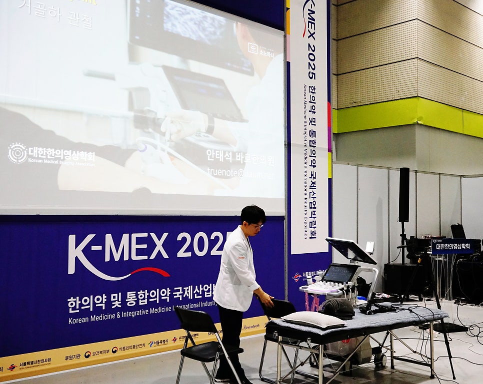 K-MEX 행사장에서 한의사가 초음파 장비와 침대 옆에 서 있고, 스크린에 초음파 관련 영상이 보이는 모습