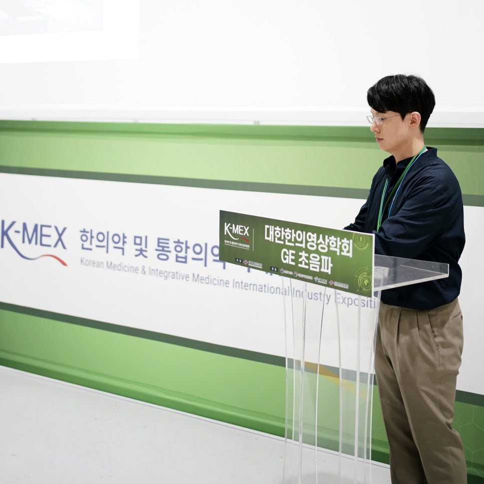 한 남성이 투명한 연단에 서 있고, 연단에는 'K-MEX 대한한의영상학회 GE 초음파'라고 적힌 표지가 놓여 있다. 뒤편에는 'K-MEX 한의약 및 통합의학'이라고 쓰인 현수막이 보인다.