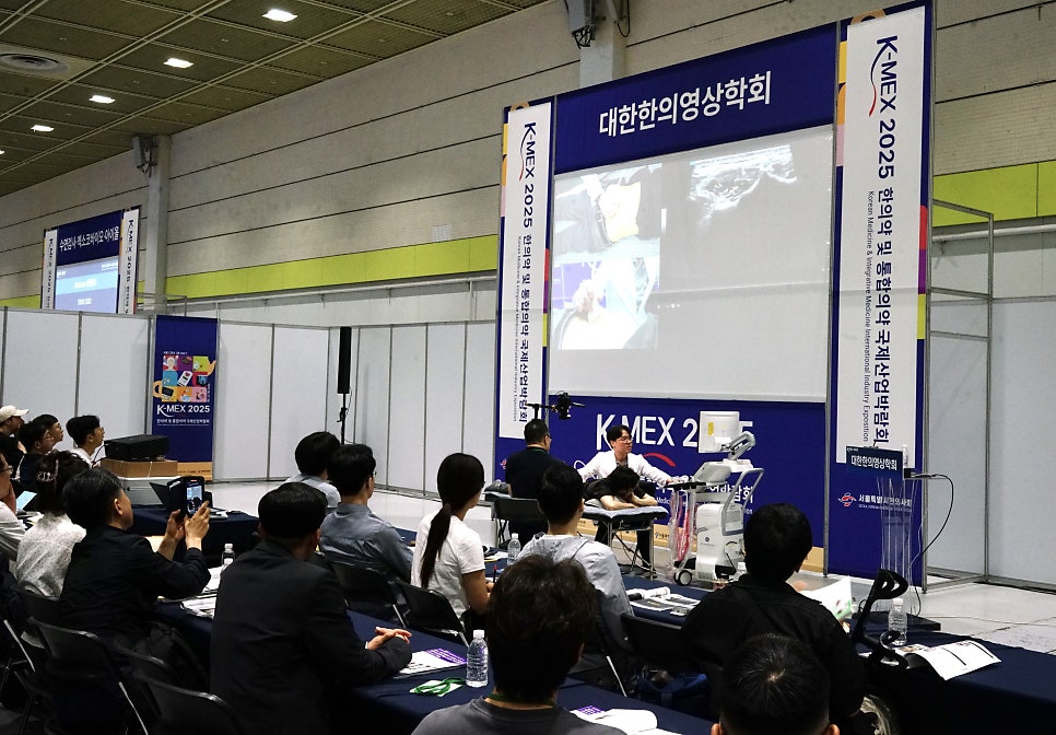 K-MEX 행사에서 강사가 초음파 기기로 시연하는 모습을 청중이 보고 있으며, 대형 스크린에 초음파 영상이 보인다.