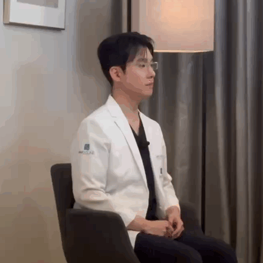 흰색 의사 가운을 입은 남성이 의자에 앉아 오른쪽을 바라보고 있다.