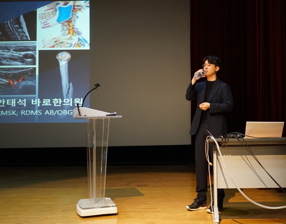 강연 중인 한의사가 물을 마시고 있으며, 뒤 스크린에는 어깨 해부학 및 초음파 이미지가 보인다.