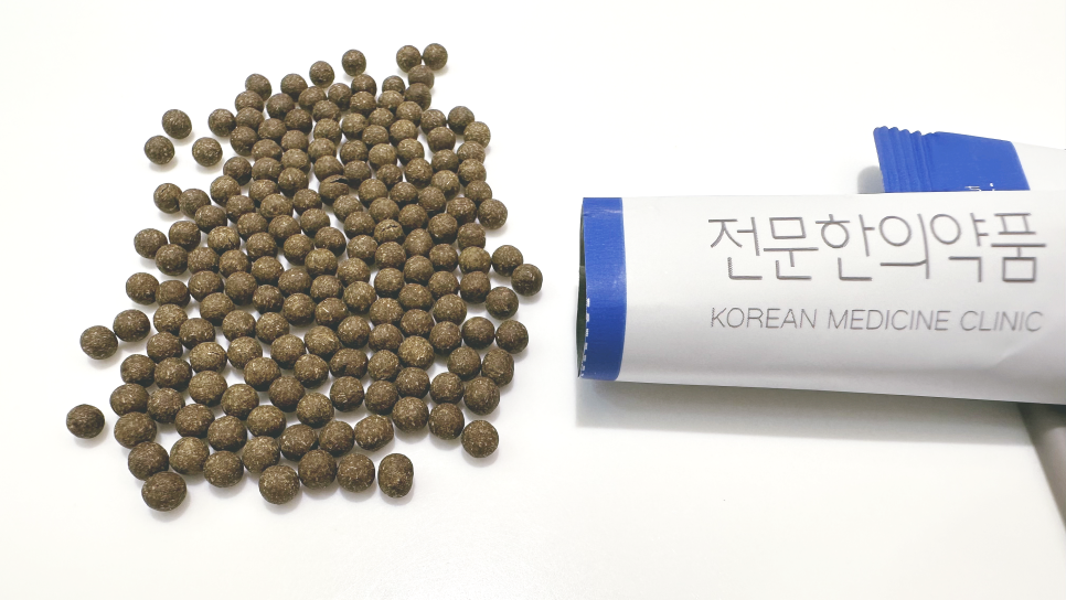 흰색 배경에 어두운 갈색 환약들이 흩어져 있고, '전문한의약품'이라고 쓰인 흰색 포장지가 옆에 있습니다.