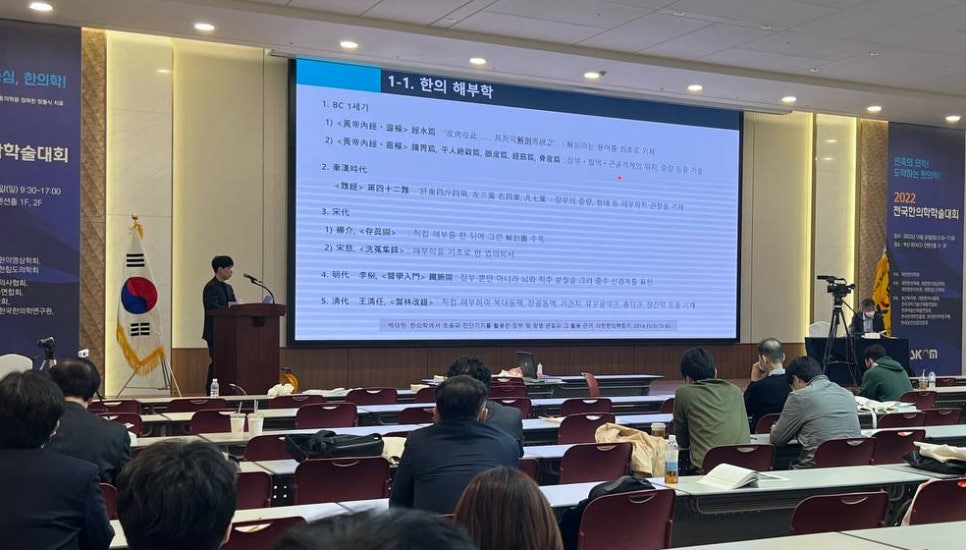 한의학 학술대회에서 연사가 '한의 해부학'이라는 제목의 발표를 하고 있으며, 청중들이 강당에 앉아 있다.