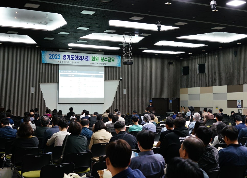 강당에 많은 사람들이 앉아 스크린에 비친 발표를 보고 있으며, '2023 경기도한의사회 회원 보수교육' 배너가 걸려 있다.