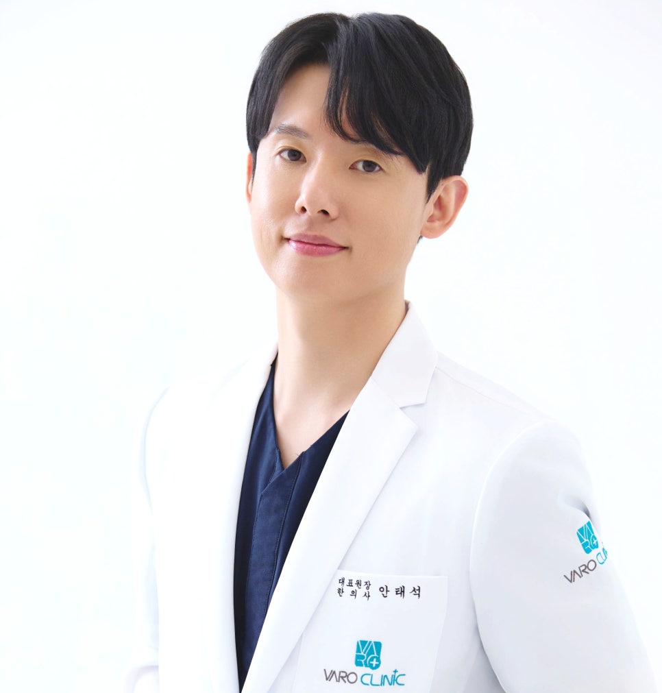 흰색 의사 가운을 입고 이름표를 단 남성이 카메라를 보며 미소 짓고 있다.