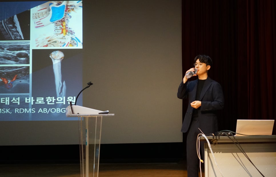 무대 위에서 정장 차림의 남자가 물을 마시고 있고, 뒤편 스크린에는 인체 해부도와 MRI, 초음파 사진 등 다양한 의료 영상이 보인다.
