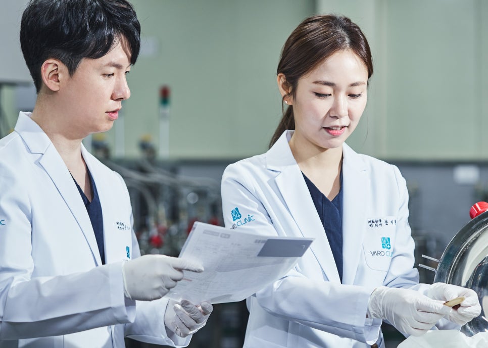흰색 가운과 장갑을 착용한 두 명의 한의사가 실험실에서 서류를 보며 작업하고 있다.