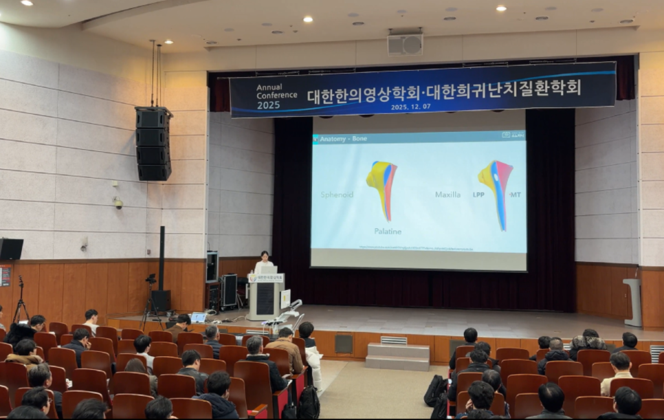 강당에서 강사가 무대 위 스크린에 뼈 해부학 이미지를 띄우고 청중에게 발표하는 모습.