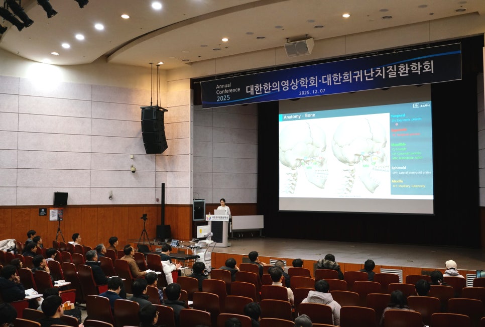 강당에서 발표자가 무대 위 스크린에 두개골 해부학 이미지를 띄우고 강연하고 있으며, 청중이 객석에 앉아 경청하고 있다.