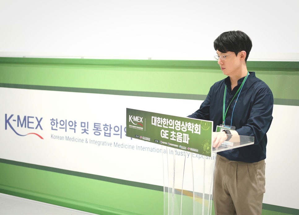 한 남성이 K-MEX 한의학 및 통합의학 배너와 대한한의영상학회 GE 초음파 표지판이 있는 연단에 서 있다.
