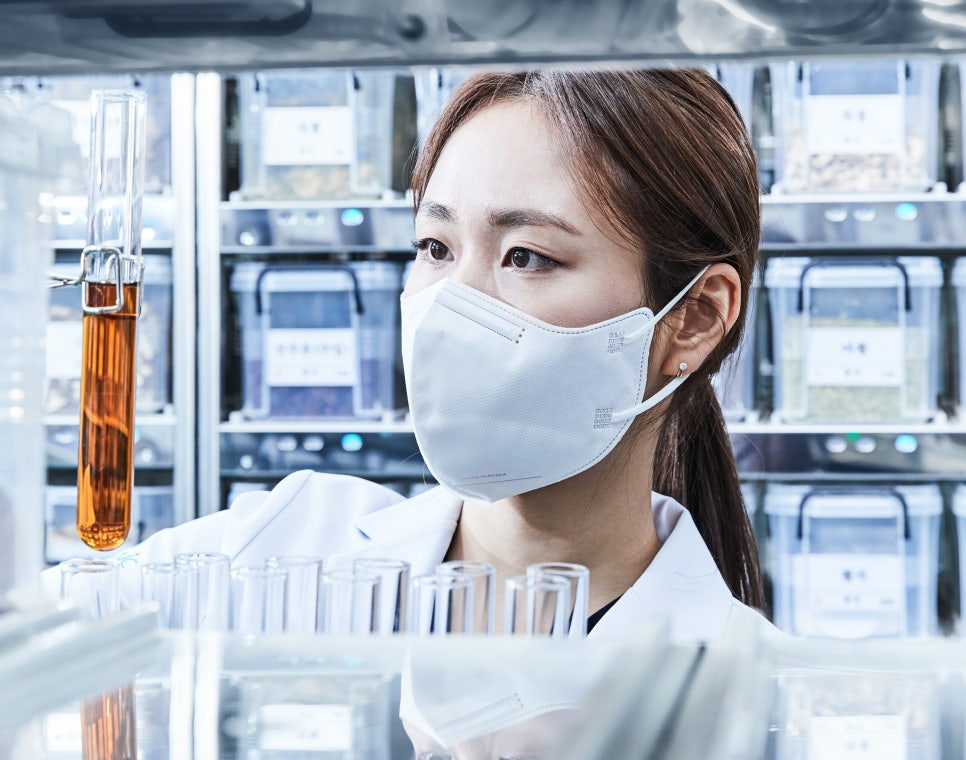 마스크를 착용한 여성이 실험실에서 시험관 속 갈색 액체를 관찰하고 있으며, 뒤편에는 한약재 용기들이 진열되어 있다.