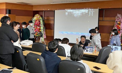 강의실에서 한의대생들이 경혈 초음파 특강을 듣고 있다. 강사가 초음파 기기를 들고 설명하며, 스크린에 초음파 영상이 투사되어 있다.