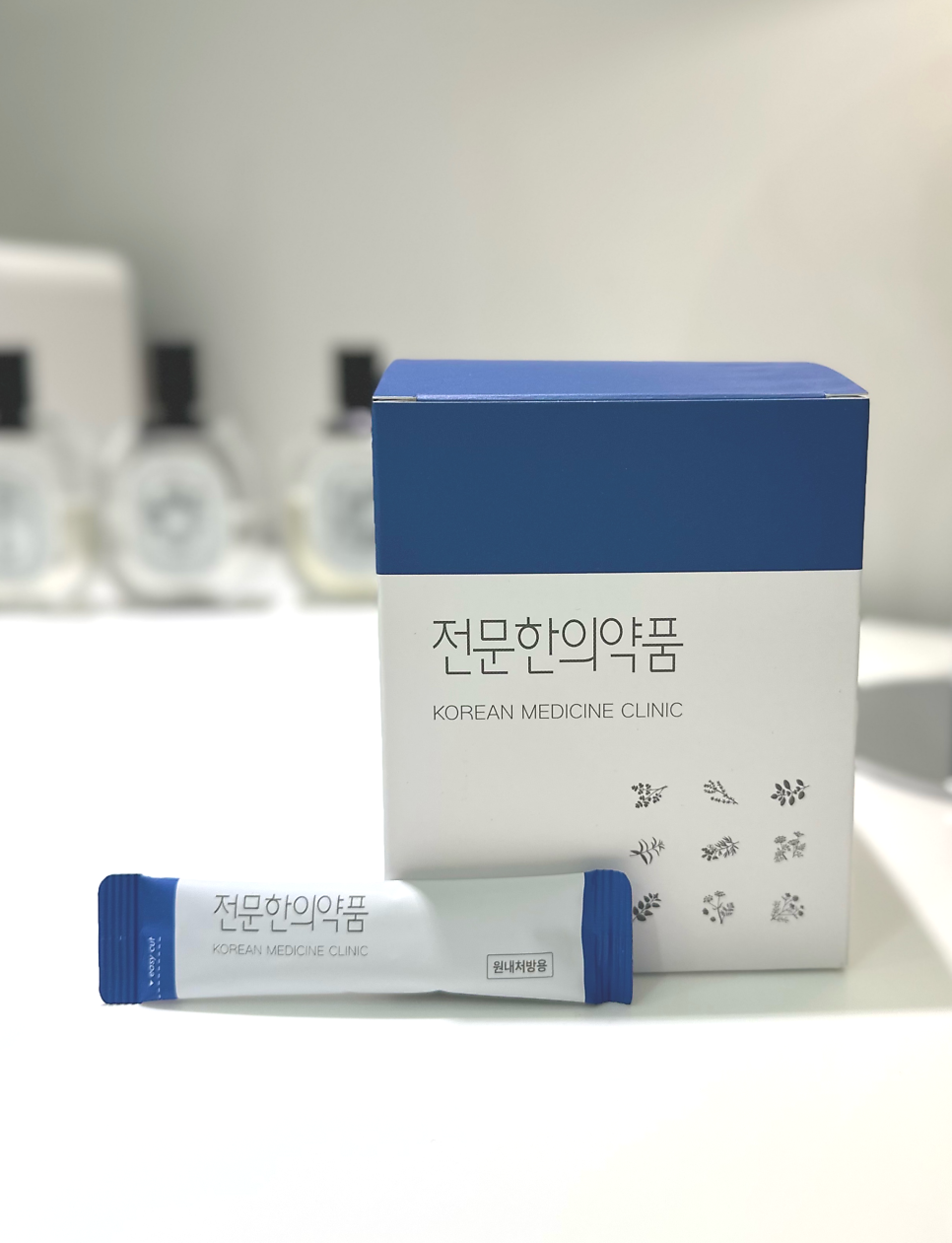 흰색과 파란색 상자 하나와 스틱형 파우치 하나가 놓여 있습니다. 상자와 파우치에는 '전문한의약품'과 'KOREAN MEDICINE CLINIC' 문구가 적혀 있으며, 파우치에는 '원내처방용' 문구도 있습니다.