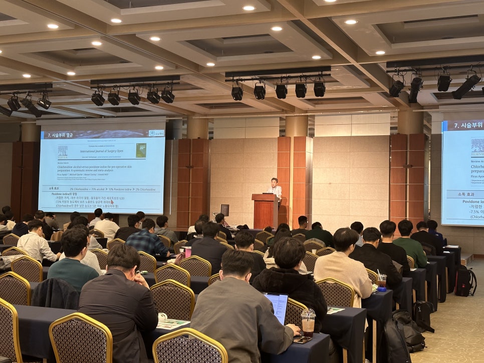 강당에서 강사가 단상에서 발표하고 있으며, 많은 참석자들이 테이블에 앉아 화면을 보고 있다.