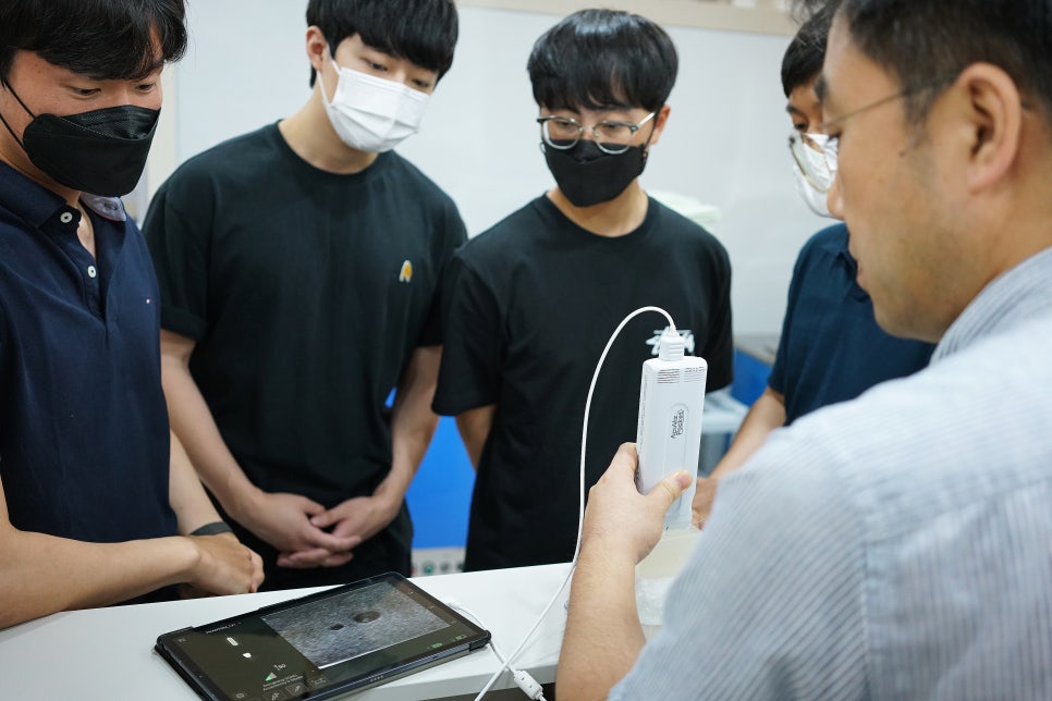 강사가 학생들에게 초음파 기기를 시연하고 있으며, 학생들이 태블릿에 보이는 초음파 영상을 보고 있다.