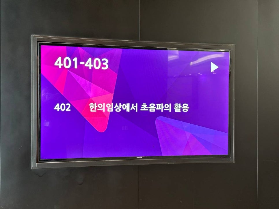 검은 벽에 걸린 디지털 화면에 '401-403', '402' 숫자와 '한의임상에서 초음파의 활용'이라는 문구가 표시되어 있다.