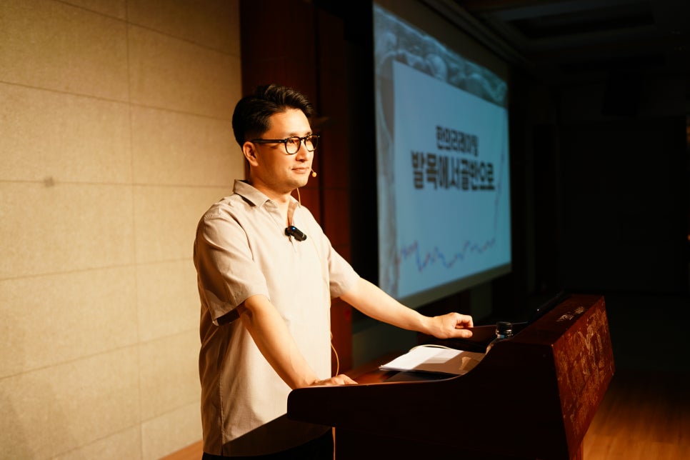 안경을 쓴 남성이 강단에서 마이크를 착용하고 발표하고 있으며, 뒤편 스크린에 글씨가 보인다.