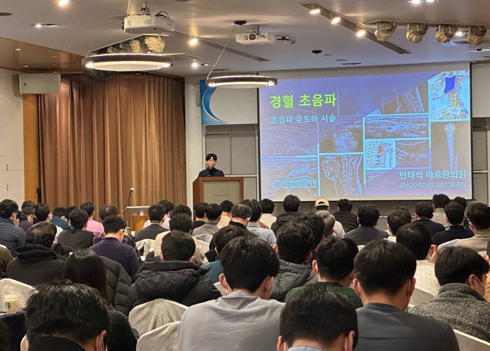강사가 연단에서 발표하고 있으며, 청중들이 앉아 경혈 초음파 및 초음파 유도 시술 강의를 듣고 있다. 스크린에는 여러 초음파 영상이 표시되어 있다.
