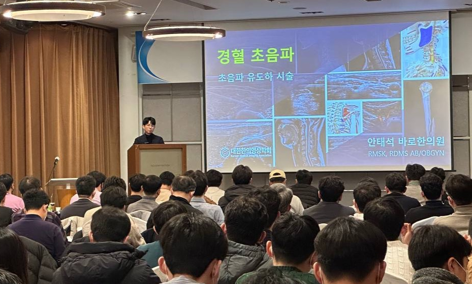 강당에서 연사가 청중에게 경혈 초음파 및 초음파 유도 시술에 대해 강연하고 있으며, 스크린에는 초음파 영상이 보인다.