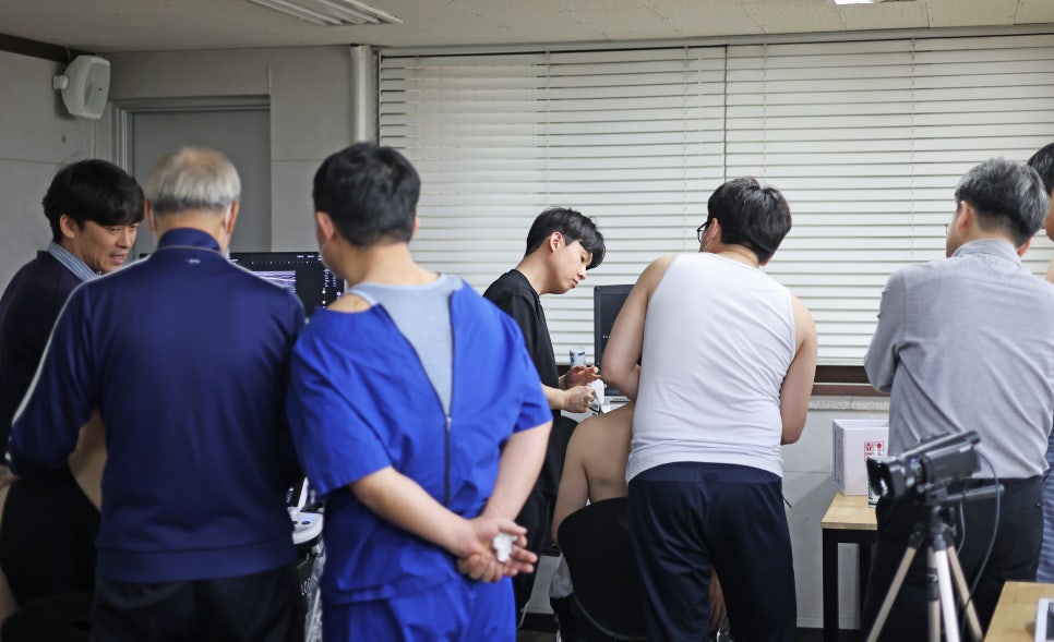 여러 명의 남성들이 모여 초음파 기기를 이용해 어깨를 검사하고 모니터를 보고 있는 모습