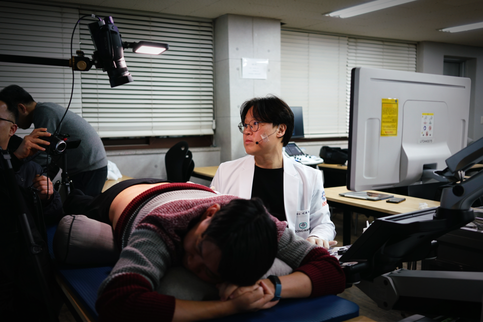 흰색 가운을 입은 남성이 침대에 엎드린 사람을 관찰하고 있으며, 뒤편에 모니터와 촬영 장비가 보인다 — 초음파 정밀 진단 | 서울 바로한의원 초음파 약침·도침·침도