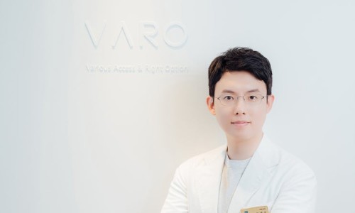 흰색 가운을 입고 안경을 쓴 남성이 'VARO' 로고가 있는 흰색 벽 앞에서 미소 짓고 있다.