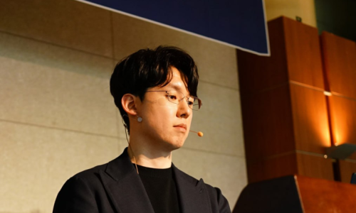 안경을 쓴 남성이 마이크를 착용하고 강단에서 아래를 보고 있다.