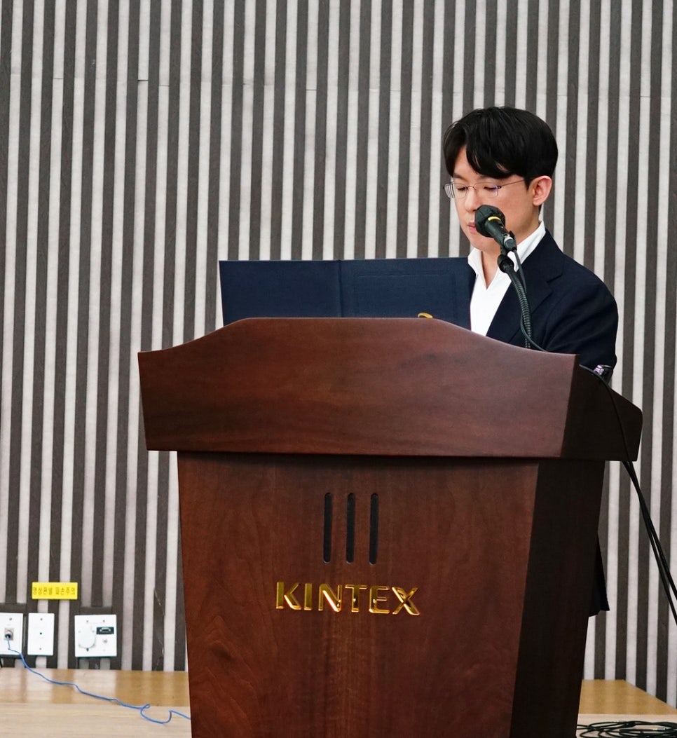 안경을 쓴 남성이 KINTEX 로고가 새겨진 나무 연단에서 마이크를 잡고 발표하는 모습