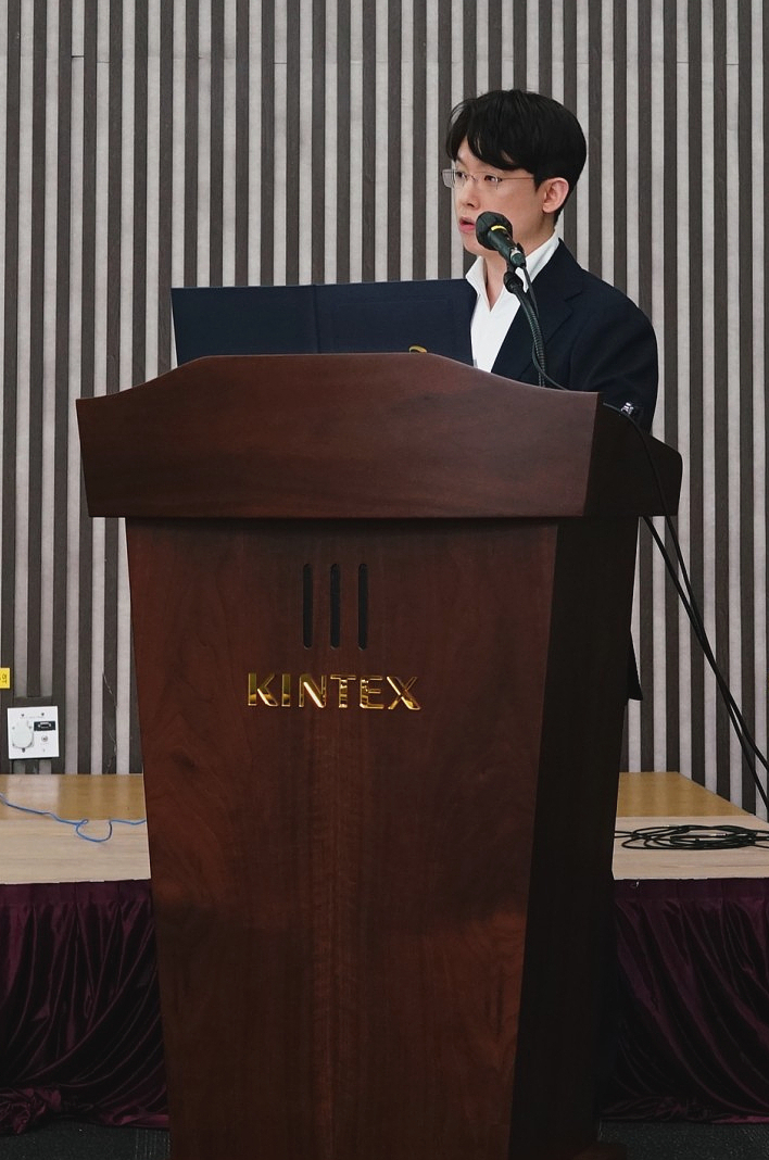 안경을 쓴 남성이 KINTEX 로고가 새겨진 나무 연단에서 마이크를 잡고 말하고 있다.