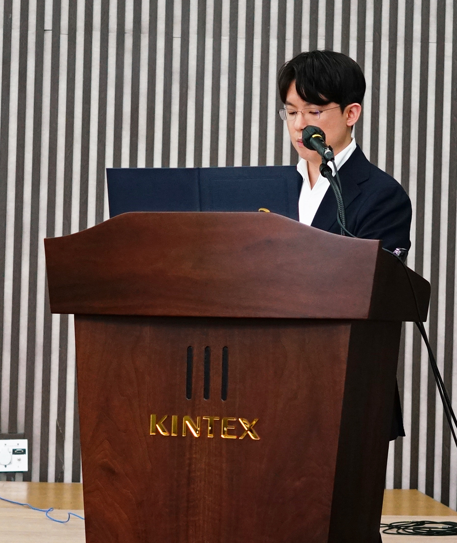 안경을 쓴 남성이 마이크 앞에서 나무 연단에 서서 발표하고 있다. 연단에는 KINTEX라고 쓰여 있다.