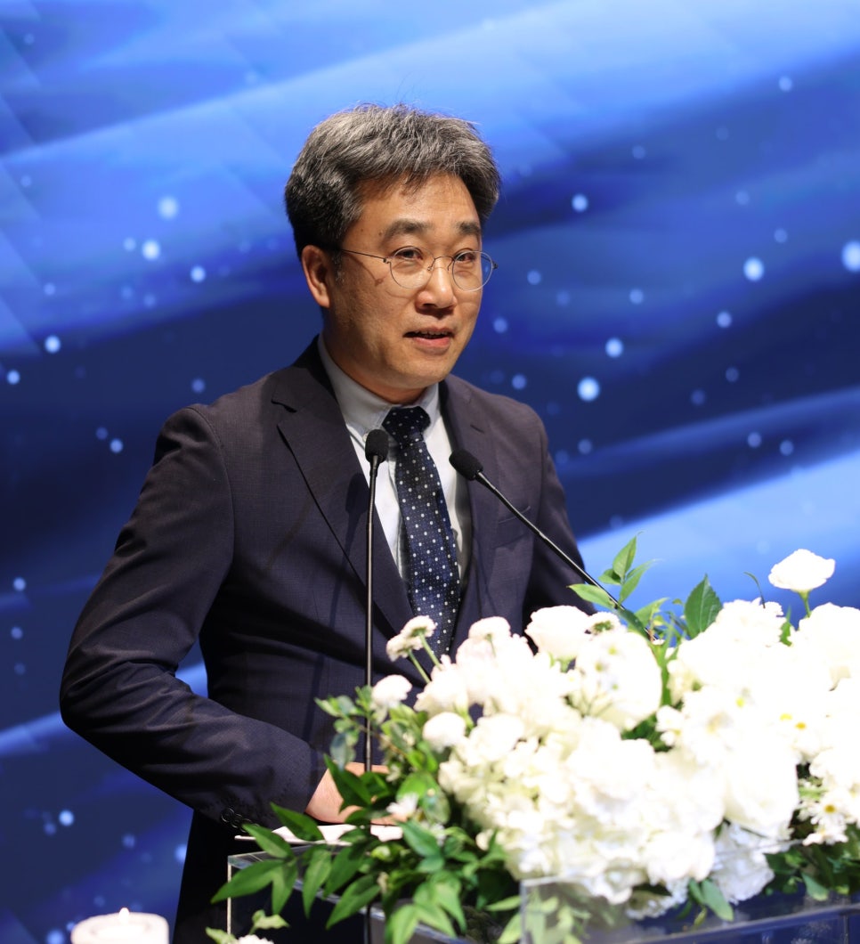 정장과 안경을 착용한 남성이 마이크가 있는 연단에서 발표하고 있으며, 연단 앞에는 흰색 꽃 장식이 놓여 있다.