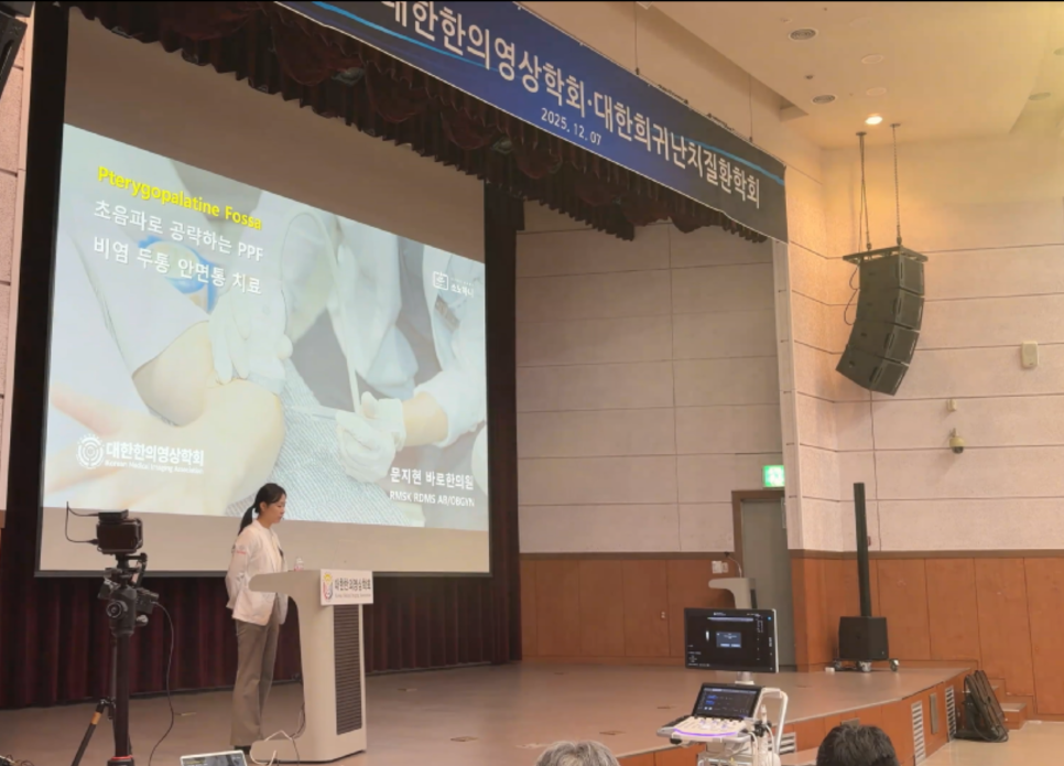 한의학회 강단에서 한 여성이 초음파 가이드 비익구개와 치료 슬라이드를 배경으로 강연하고 있다.