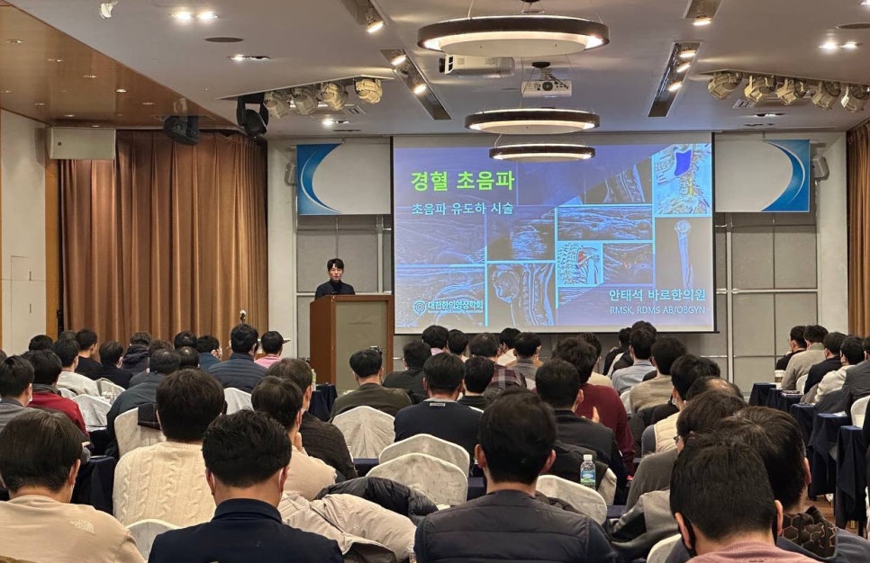 강사가 강연장에서 청중에게 초음파 유도 시술에 대해 발표하고 있다. 스크린에는 '경혈 초음파'와 '초음파 유도하 시술'이라는 문구와 관련 이미지가 보인다.
