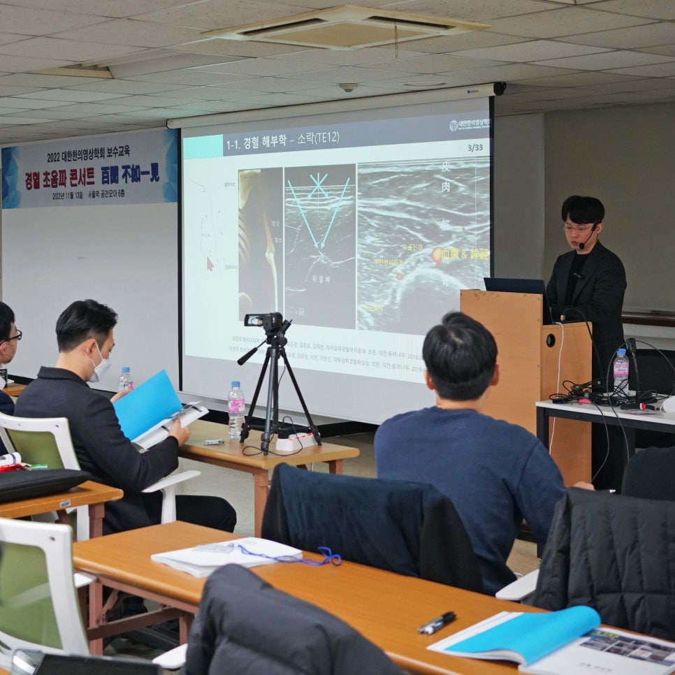 강사가 스크린에 경혈 해부학 및 초음파 이미지를 보여주며 발표하고 있고, 참석자들이 앉아서 보고 있는 세미나 모습.