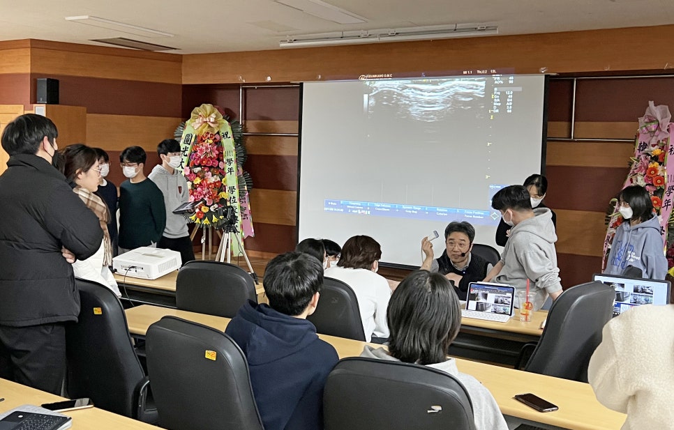 강의실에서 강사가 학생들에게 경혈 초음파 특강을 진행하며 스크린에 초음파 영상이 보이는 모습