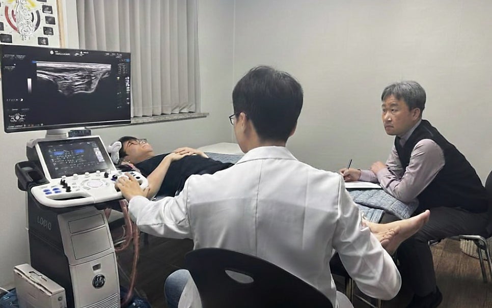 한의사가 환자의 다리에 초음파 검사를 하고 있으며, 초음파 화면이 보이고 다른 한의사가 이를 지켜보고 있다.