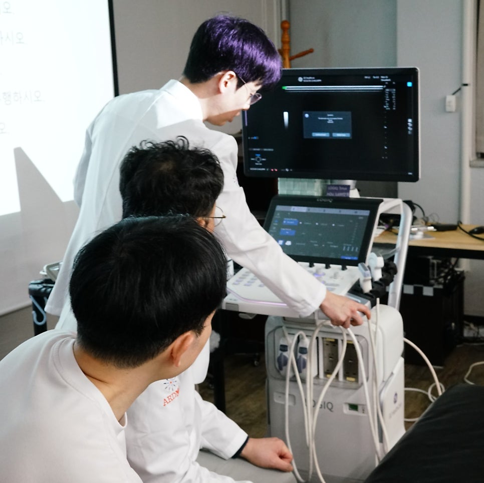 흰색 가운을 입은 한 사람이 초음파 기기를 조작하고 있고, 다른 두 사람이 이를 지켜보고 있는 모습.