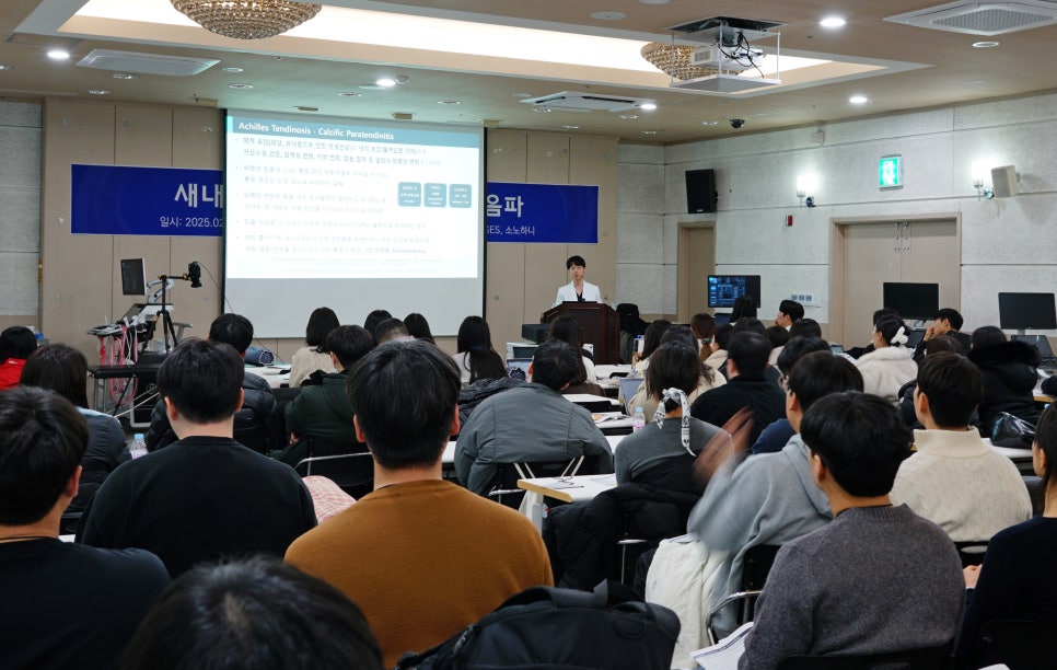 스크린에 아킬레스건염 관련 내용이 표시된 강의실에서 강사가 청중에게 발표하고 있다.