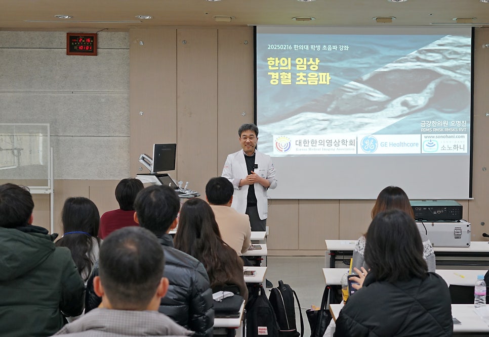 한의사가 강단에서 경혈 초음파 강의를 하고 있으며, 여러 명이 앉아 강의를 듣고 있는 모습.