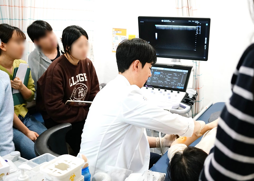 한의사가 환자의 팔에 초음파를 보며 시술하고 있으며, 다른 사람들이 이를 지켜보고 있다.