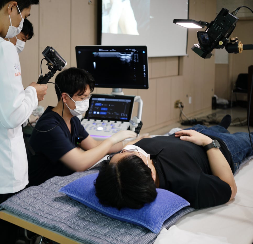 한의사가 환자에게 초음파 기기를 사용하고 있으며, 모니터에 초음파 영상이 보인다. 주변에 다른 사람들이 지켜보고 있다.