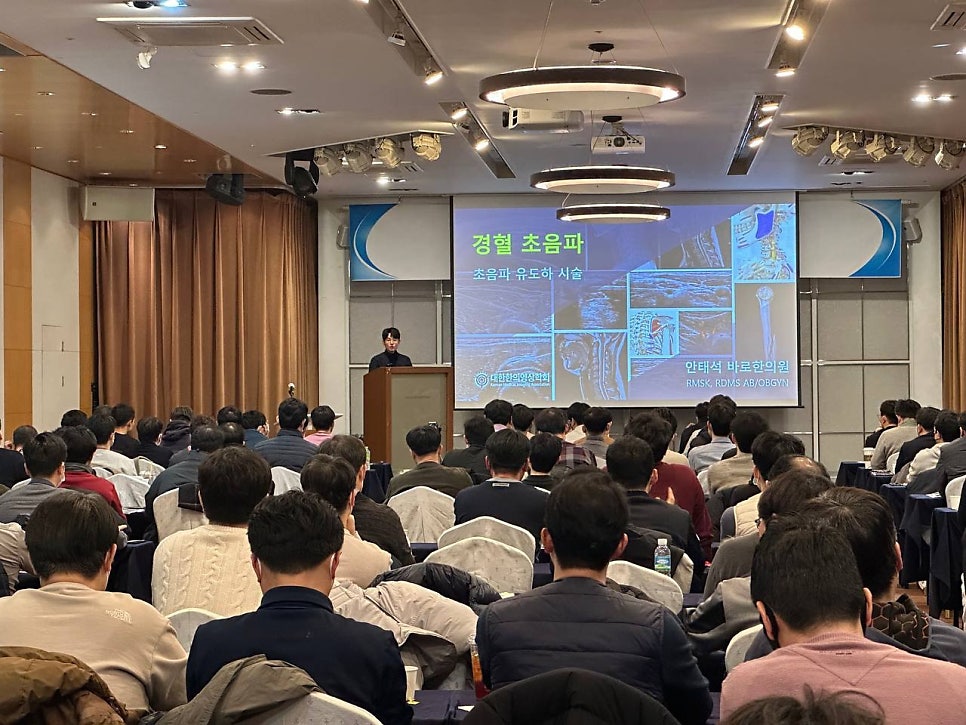 강당에서 강사가 정형 초음파와 초음파 유도 시술에 대해 발표하고 청중이 앉아 있는 모습.