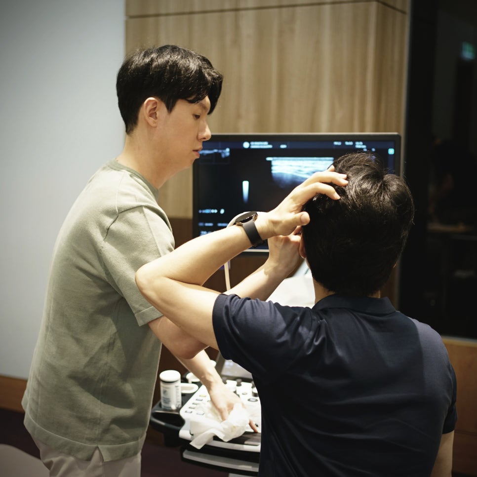한 남자가 초음파 기기 앞에서 서서 초음파 화면을 보고 있고, 다른 남자는 앉아서 화면을 보고 있다.