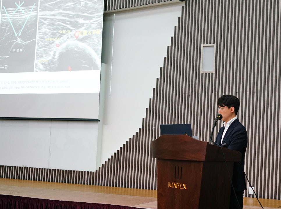 킨텍스 로고가 있는 강단에서 한 남성이 발표하고 있으며, 뒤 스크린에는 초음파 영상과 의료 관련 자료가 표시되어 있다.