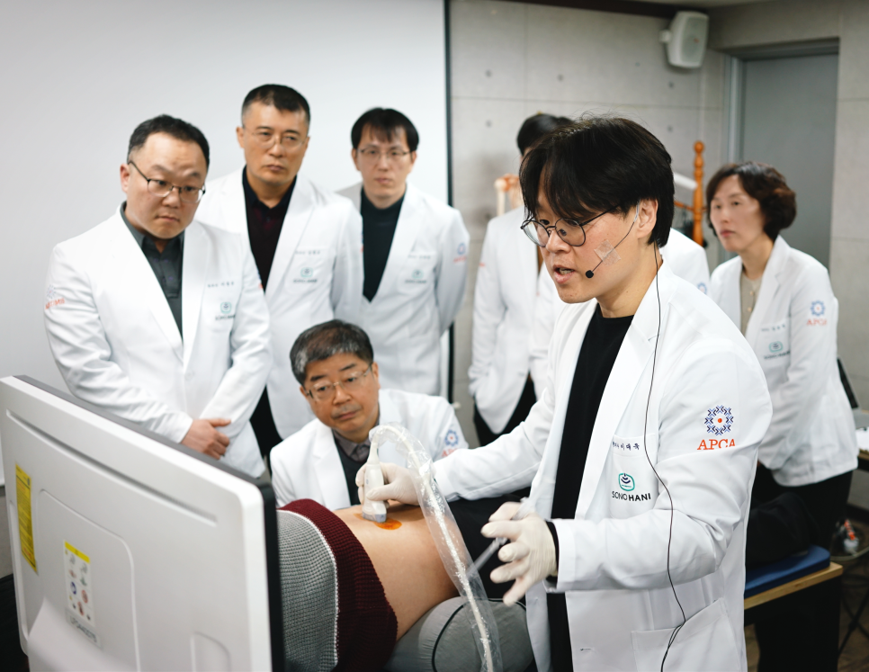 흰 가운을 입은 강사가 환자의 허리에 초음파 기기를 사용하며 설명하고, 여러 사람이 이를 지켜보고 있다 — 초음파 정밀 진단 | 서울 바로한의원 초음파 약침·도침·침도
