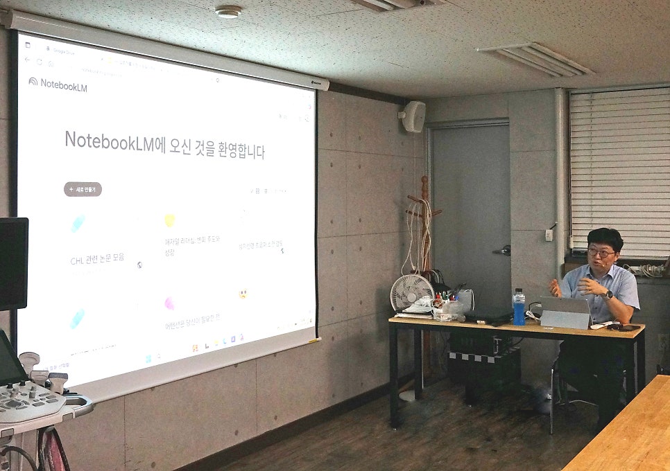 강사가 노트북LM 화면을 띄운 프로젝터 앞에서 발표하고 있으며, 왼쪽에 초음파 기기가 놓여 있다.