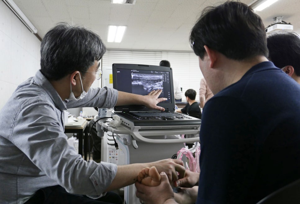 마스크를 쓴 사람이 초음파 기기 화면의 초음파 영상을 가리키고 있고, 다른 사람의 발에 초음파 탐촉자가 놓여 있다.