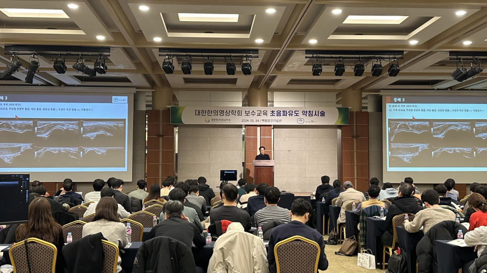 강당에서 강사가 연단에 서서 청중에게 강연하고 있으며, 양쪽 스크린에는 초음파 영상이 보인다.