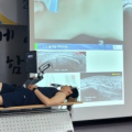 한 사람이 테이블에 누워 있고, 뒤쪽 스크린에 초음파 영상과 시술 장면이 보인다.