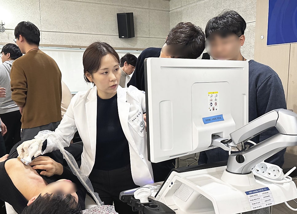 한의사가 환자의 상체에 초음파 기기를 대고 모니터를 보며 진료하고 있으며, 주변 사람들이 이를 지켜보고 있습니다.